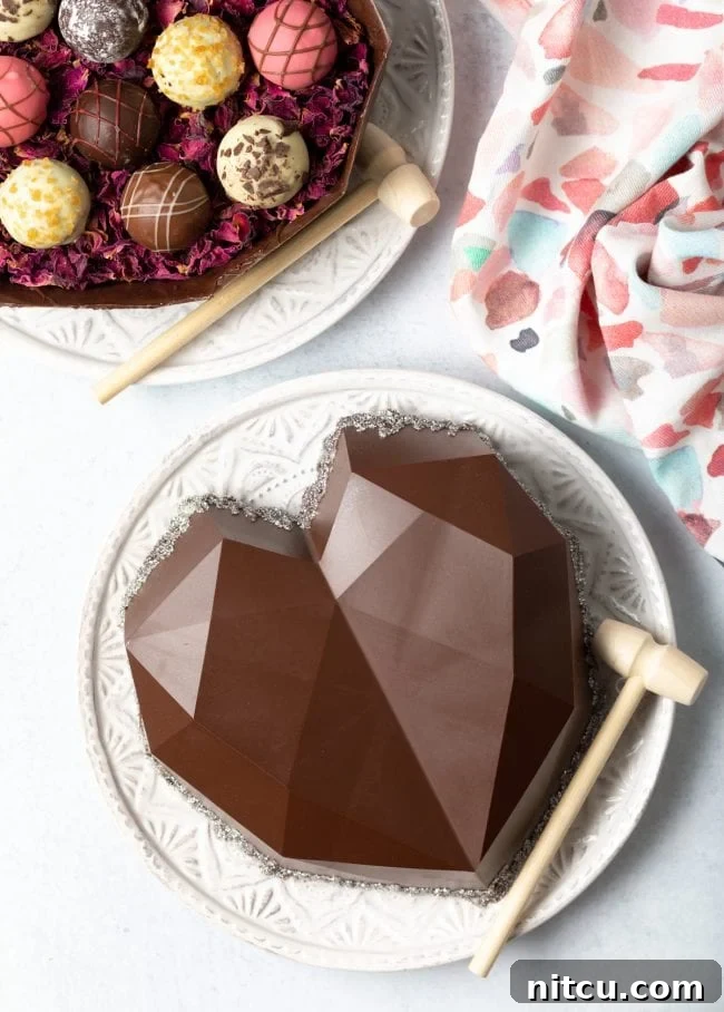 A top-down view showcases a heart-shaped chocolate box on a white plate, complete with a small hammer. The open bottom half of the box, filled with truffles, is visible in the upper left corner.