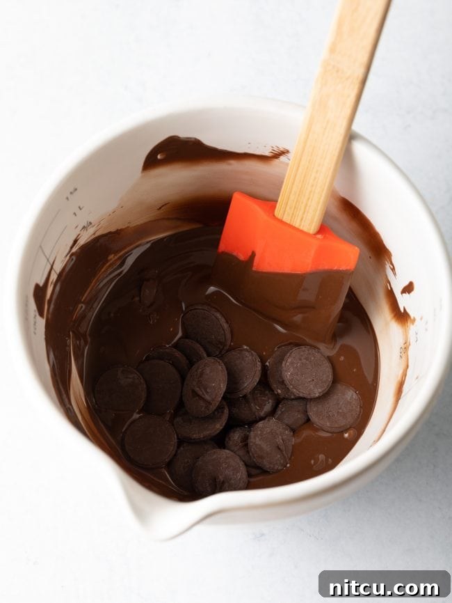 Glossy chocolate wafers are captured in the process of melting within a white measuring cup, stirred by a clean spatula, ready for molding.