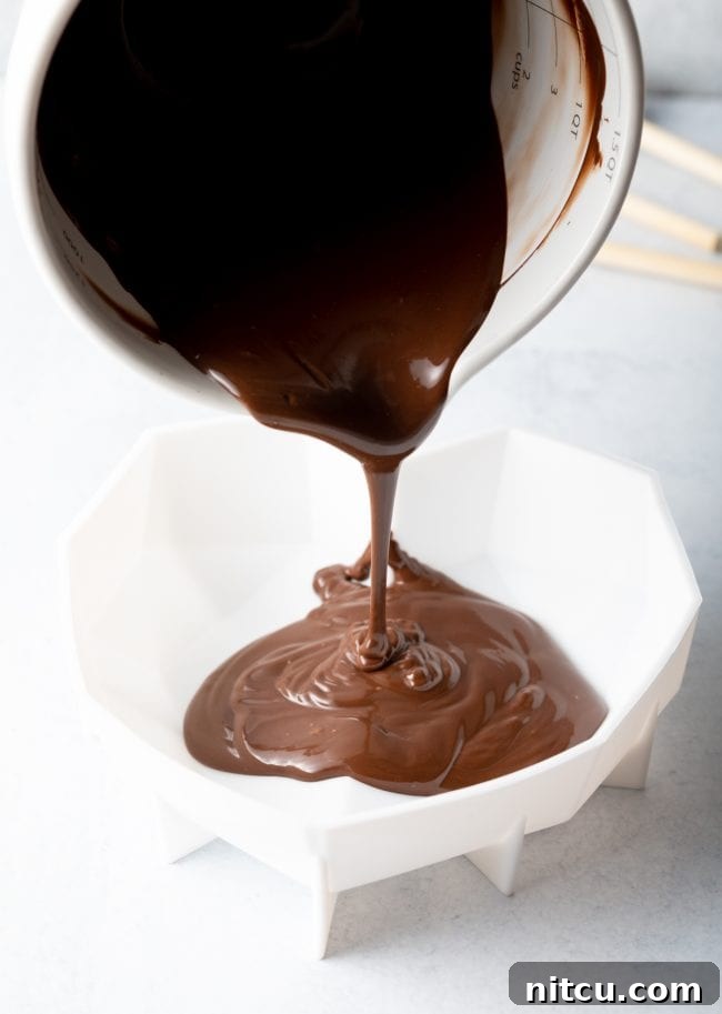 Melted chocolate is expertly poured from a white measuring cup into a flexible white silicone heart shell mold, ensuring a perfect coating.