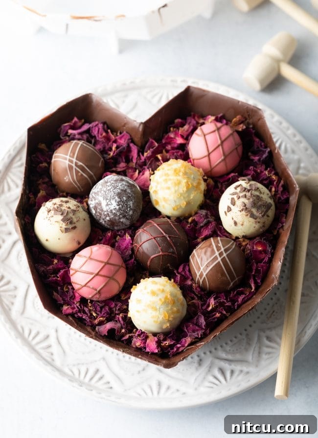 The bottom half of a breakable chocolate heart rests on a white plate, elegantly filled with delicate edible rose petals and an assortment of gourmet truffles.