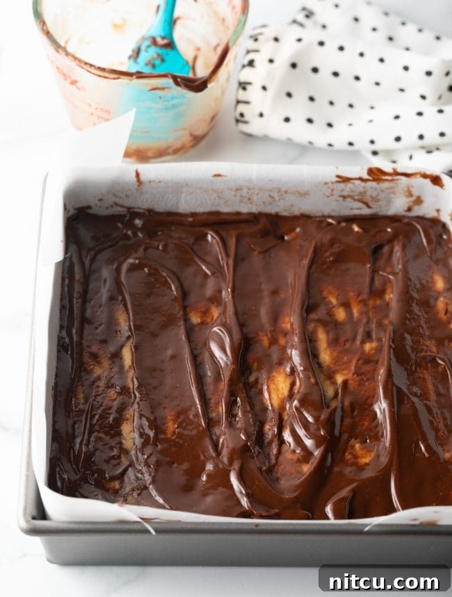 Top-down view of melted chocolate fudge topping being added and swirled onto the peanut butter base in a baking pan.