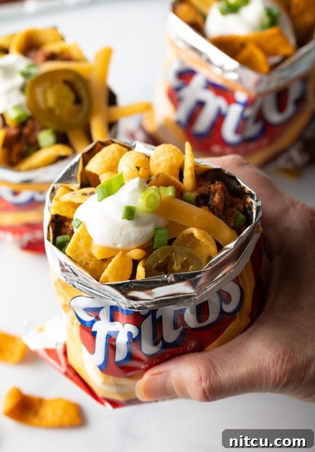 A hand holding an open bag of Fritos corn chips filled with a complete Frito chili pie, topped with chili, cheese, and other garnishes.