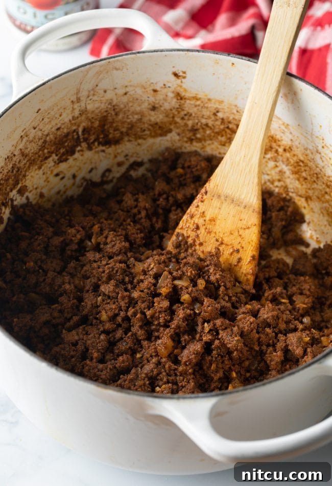 Lean ground beef being browned in a large pot, stirred with a wooden spoon, alongside diced onions and garlic.