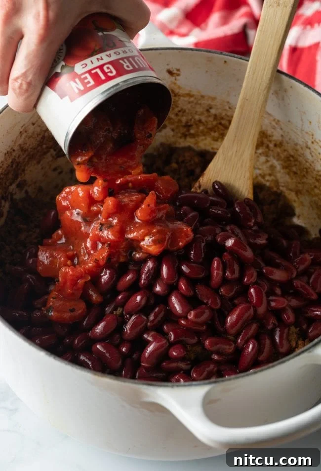 A hand adding canned diced tomatoes to a pot containing kidney beans and browned beef, stirred with a wooden spoon.