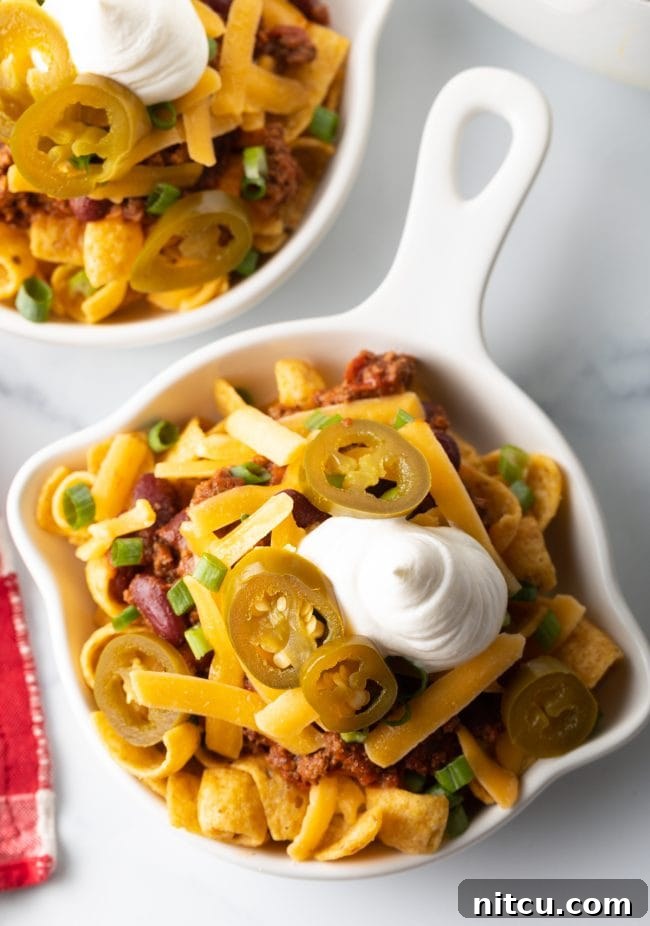 A top-down view of a finished Frito chili pie in a shallow white dish, adorned with chili, cheese, scallions, jalapenos, and sour cream.
