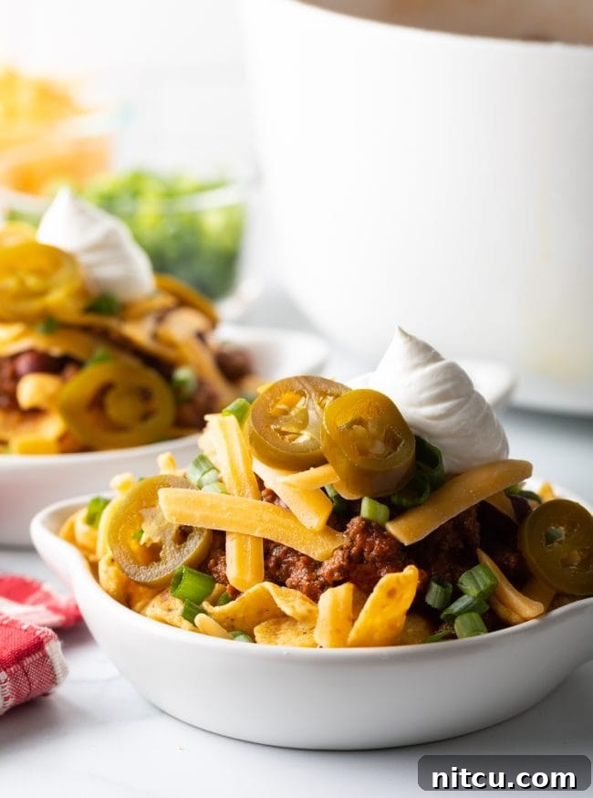 A side view of a white shallow dish brimming with Frito chili pie, showcasing layers of beef chili, crunchy corn chips, shredded cheese, chopped scallions, tangy jalapeno slices, and a creamy dollop of sour cream.