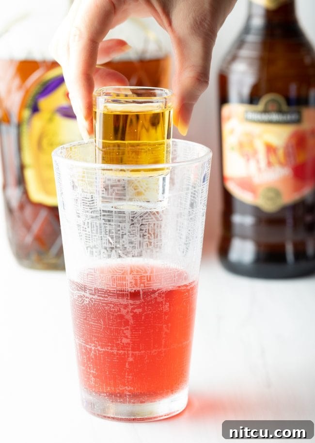 hand holding shot of red bull above the glass of whisky, cranberry juice, and peach schnapps
