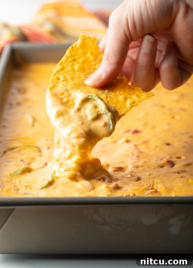 Hand scooping creamy smoked queso dip with a jalapeño garnish from a large metal pan, ready for serving.