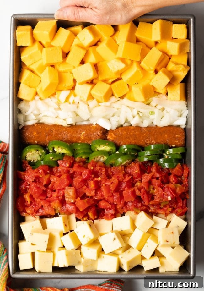 Top view of a metal pan filled with rows of pepper jack cheese, Rotel, sliced jalapeños, Mexican chorizo, diced onions, and Velveeta cubes, ready for smoking.
