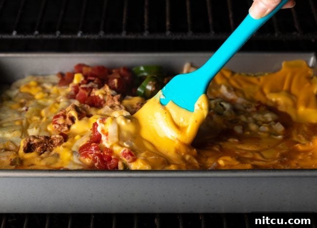 A metal pan of smoked queso dip being stirred with a spatula inside a smoker, creating a creamy texture.