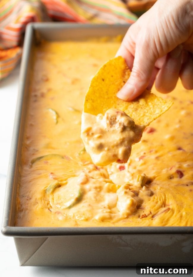 A hand with a tortilla chip scoops rich Mexican chorizo smoked queso cheese dip from a metal pan, highlighting its creamy texture.