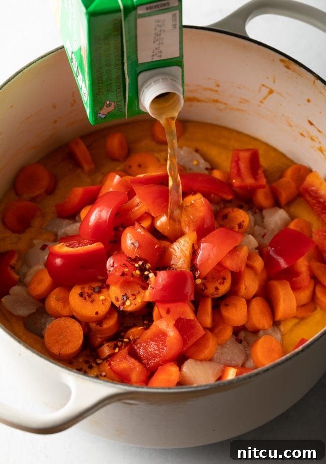 Chicken broth being poured over bell peppers, carrots, chicken, and the creamy Maafe base in a pot.