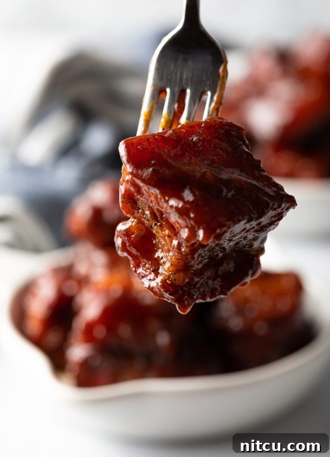 A fork holding a perfectly glazed cube of smoked pork belly burnt ends, ready to be eaten.