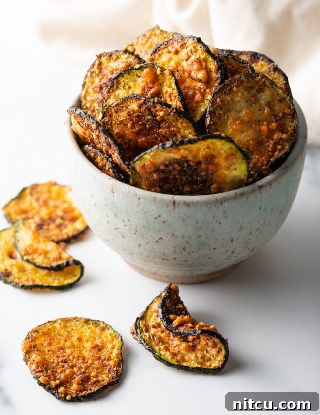 Full view of a speckled bowl overflowing with parmesan zucchini crisps and chips scattered around on a wooden surface.