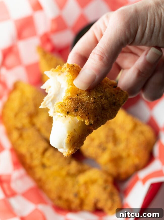 A hand presenting a piece of perfectly fried cornmeal fish towards the camera, showcasing its golden, crispy crust.