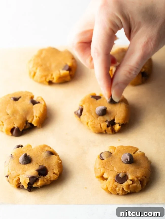 A hand carefully pressing extra chocolate chips onto flattened raw cookie dough on parchment paper, enhancing its visual appeal.
