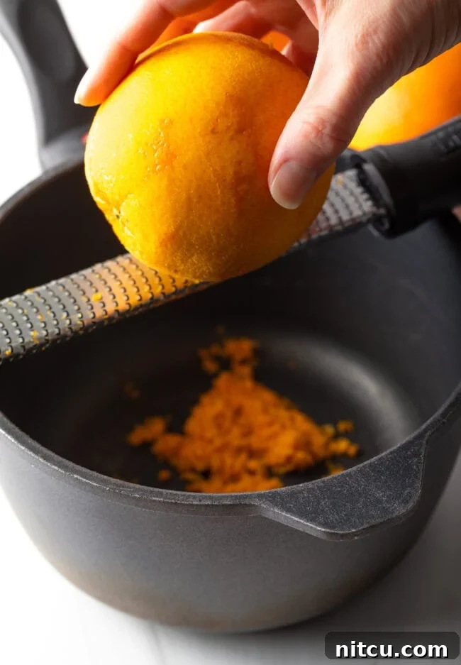 Hand zesting an orange above a bowl. A hand skillfully zesting a fresh orange directly over a bowl, capturing the fine, aromatic zest. This image emphasizes the importance of fresh citrus zest in the recipe and the technique for gathering it.