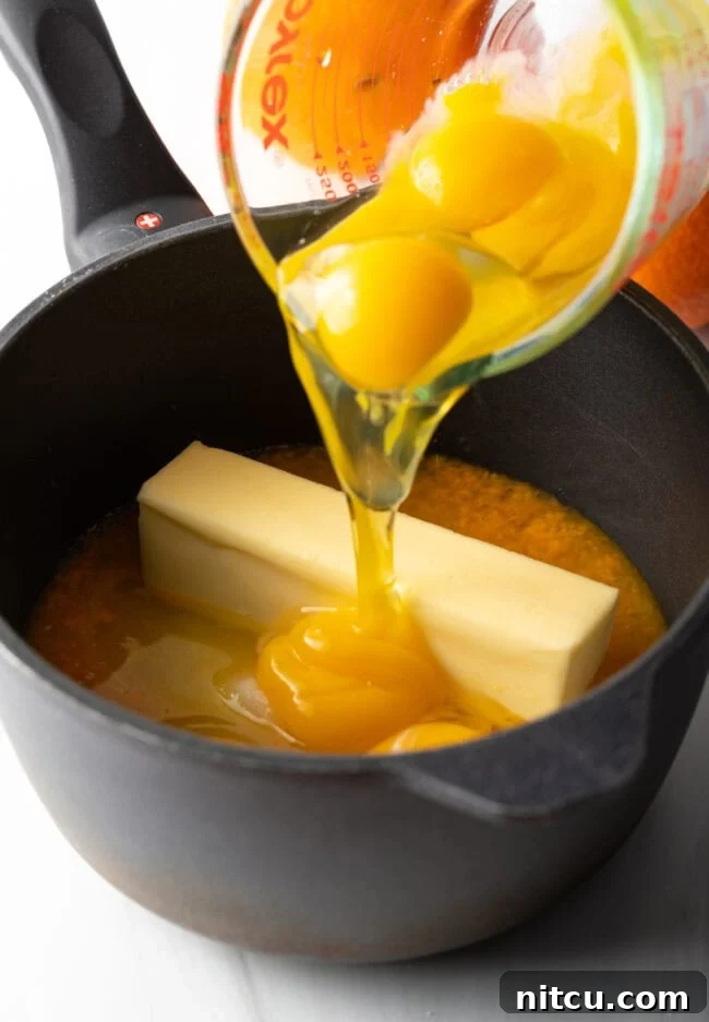 Glass measuring bowl adding egg yolks and butter to the pot. A glass measuring bowl being used to add egg yolks and cubes of butter into a saucepan. This step illustrates the careful incorporation of key ingredients that will give the orange curd its rich, creamy texture.