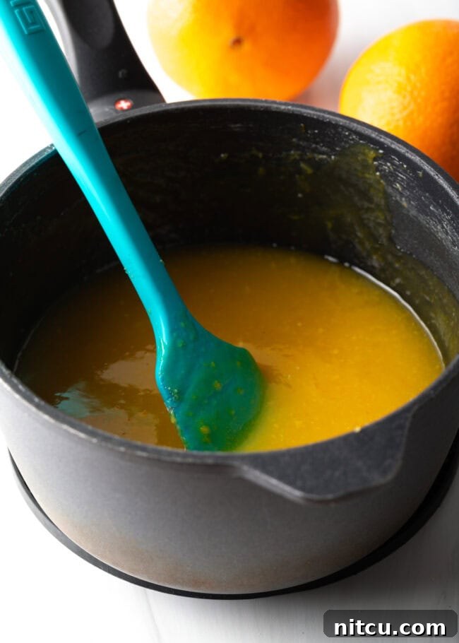 Pot cooking orange curd with blue rubber spatula stirring. A blue rubber spatula actively stirring vibrant orange curd in a pot as it cooks on the stovetop. This action shot emphasizes the importance of continuous stirring to achieve a smooth, lump-free curd, preventing it from sticking or burning.
