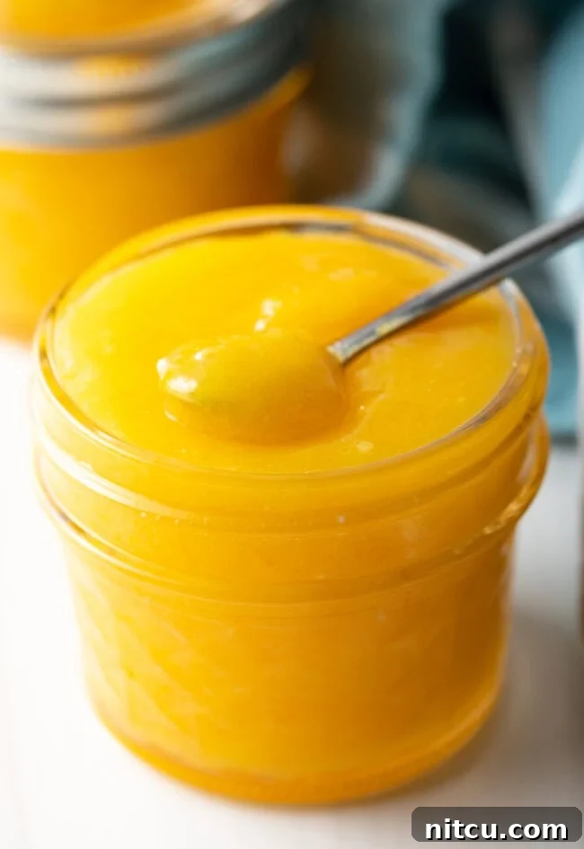 Finished jar of homemade orange curd with little spoon. A charming shot of a glass jar filled with luscious homemade orange curd, accompanied by a small spoon ready for tasting. The inviting texture and vibrant color signal a fresh, delicious spread, perfect for any occasion.