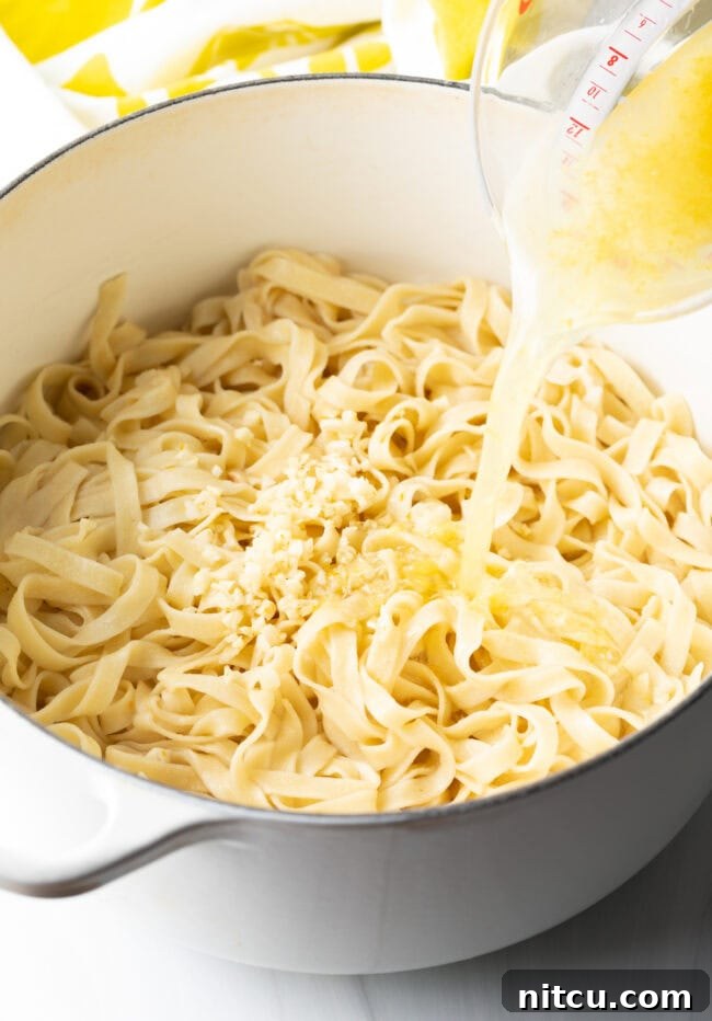 Glass measuring cup adding lemon juice, water, and minced garlic to the pot of fettuccine noodles.