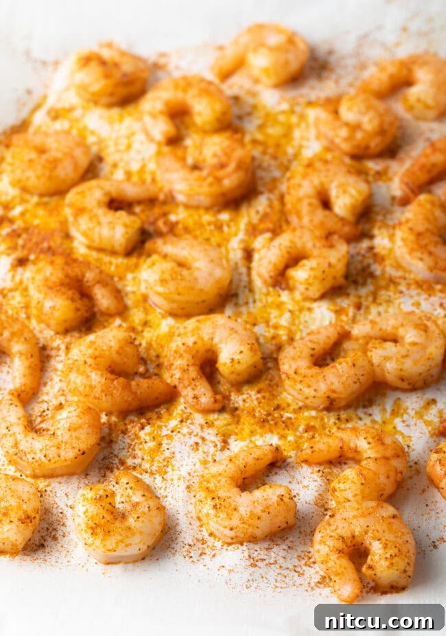 Oven-baked seasoned shrimp spread in a single layer on a parchment-lined baking sheet, showing their pink, curled shape.