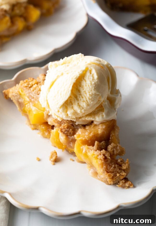 A close-up shot of a slice of peach crumble pie, served on a plate with a scoop of melting vanilla ice cream on top.