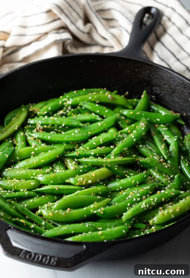 Top down view of vibrant green sugar snap peas stir-frying in a skillet with sesame seeds.