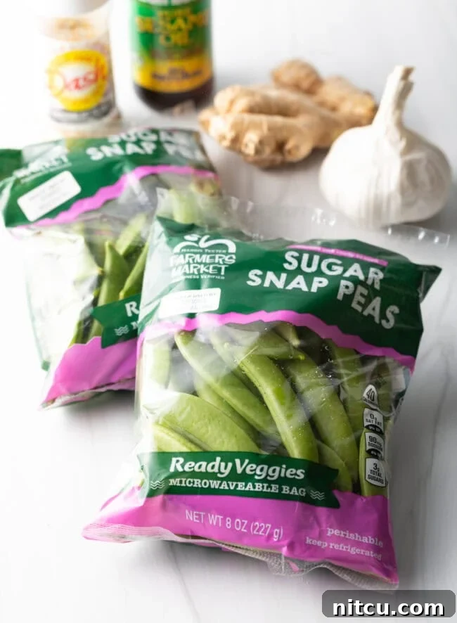 Fresh ingredients laid out: a bag of sugar snap peas, a head of garlic, a piece of ginger, and small bowls of seasonings in the background.