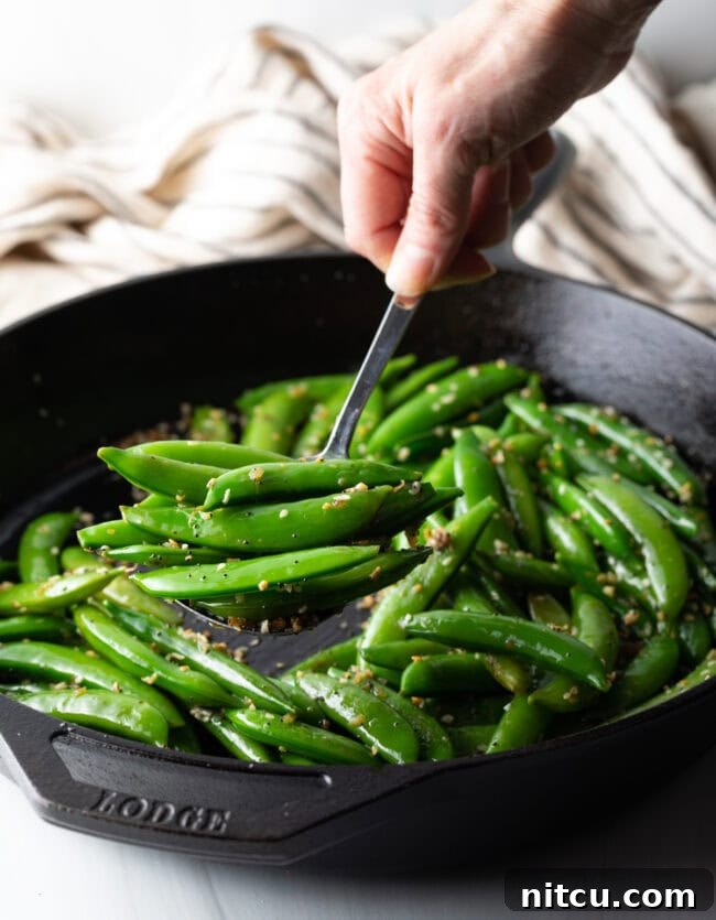 A hand stirring bright green sugar snap peas in a large metal skillet with a metal spoon, steam rising.