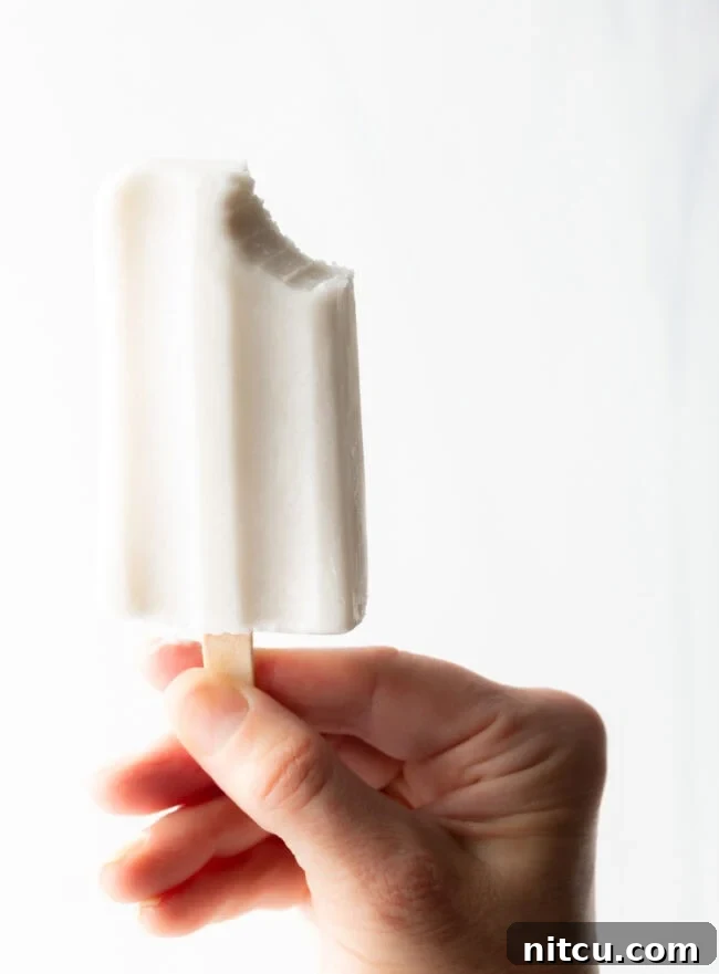 A hand holding a coconut milk popsicle with a bite taken, revealing its creamy texture against a blurred background.