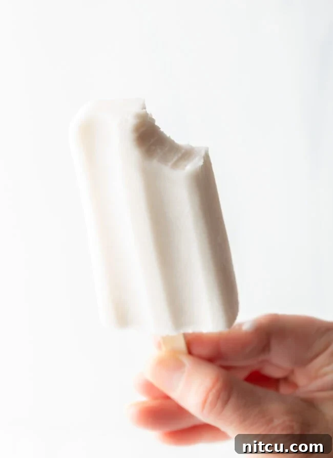 A hand holding a perfectly creamy coconut milk popsicle with a small bite taken out, highlighting its smooth texture.