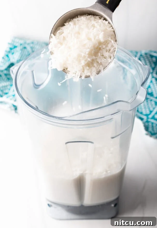 Shredded coconut being added to a blender with creamy coconut milk, sugar, vanilla, and salt for the popsicle mixture.