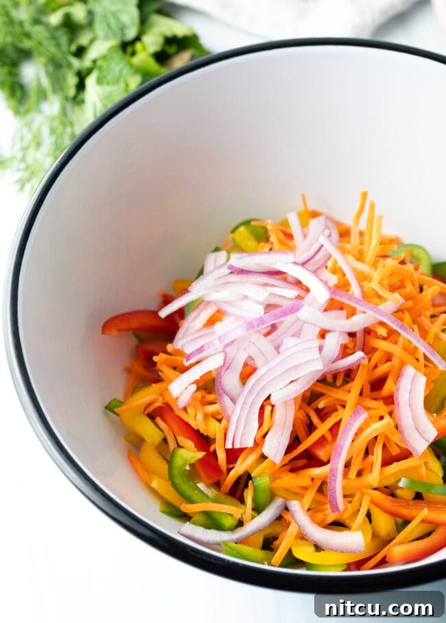Sliced bell peppers, shredded carrots, and thinly sliced red onion in a large mixing bowl, ready for herbs and dressing.