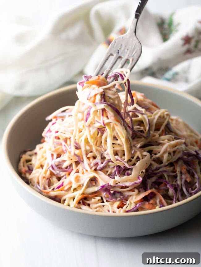 A fork taking a bite of creamy coleslaw from a white bowl, highlighting the delicious texture.