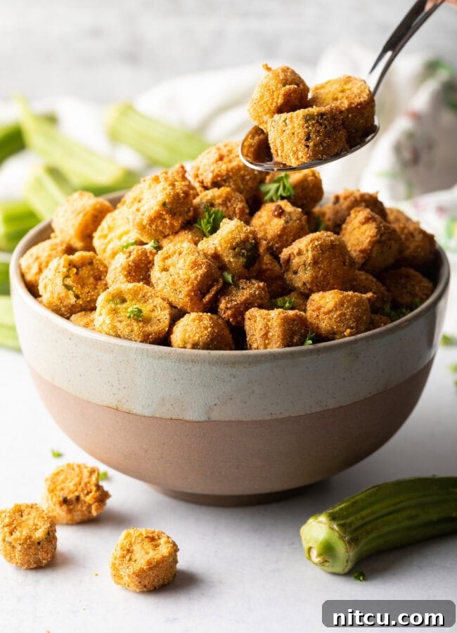 A spoon carefully adding golden pieces of Southern fried okra into a serving bowl.
