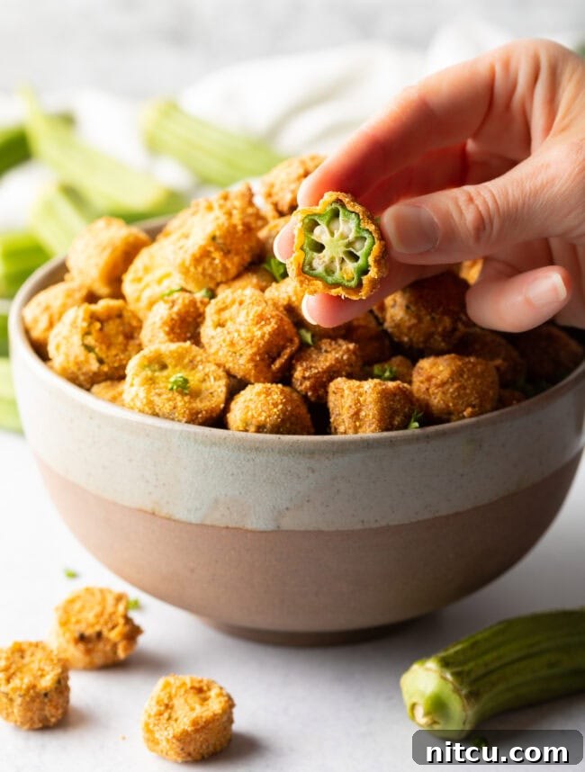 A hand holding a perfectly sliced and breaded piece of fried okra up to the camera, showcasing its golden exterior.