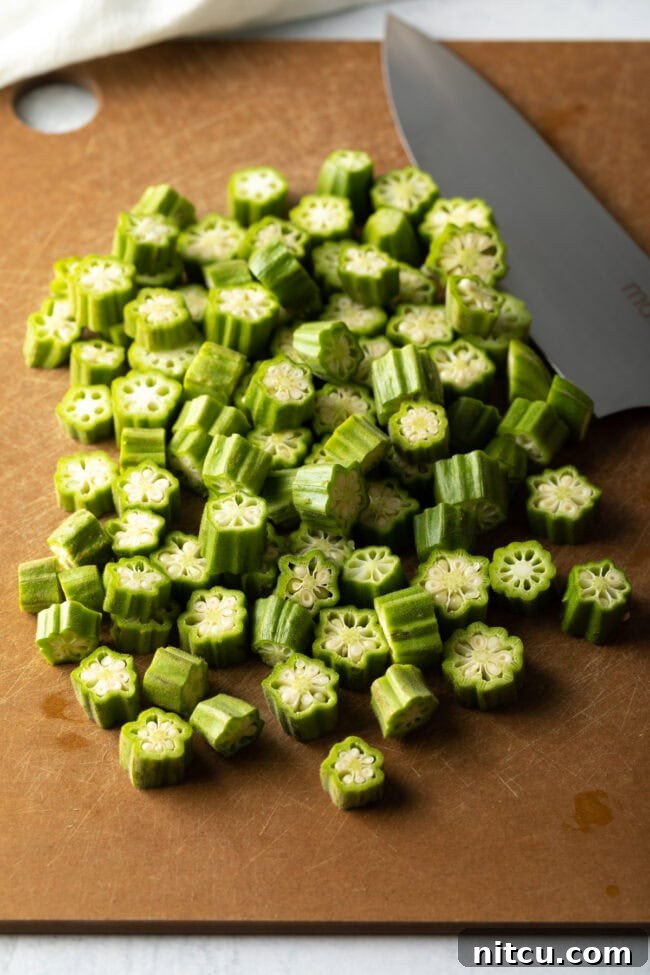Fresh okra being sliced into uniform rounds on a wooden cutting board with a chef's knife.