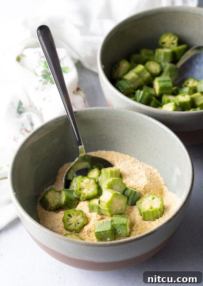 Okra slices being added to a bowl with cornmeal breading mixture, ensuring full coverage.