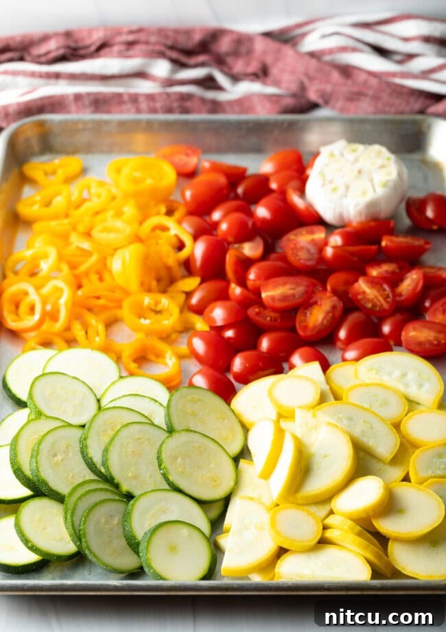 Sheet pan with piles of sliced zucchini, summer squash, tomatoes, peppers, and garlic, ready for roasting.