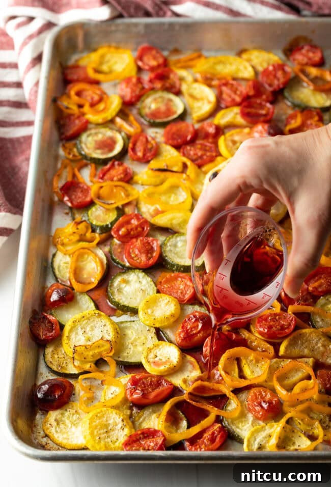 Hand pouring red wine over roasted summer vegetables on a sheet pan.