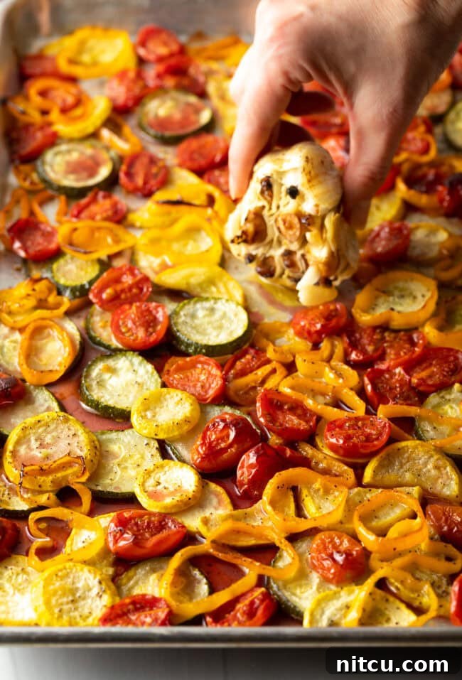 Hand squeezing out soft roasted garlic cloves over the pan of roasted vegetables.