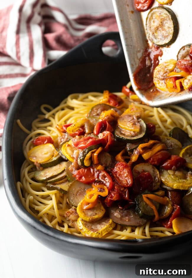 Pouring roasted vegetables and sauce over the spaghetti in a large bowl.