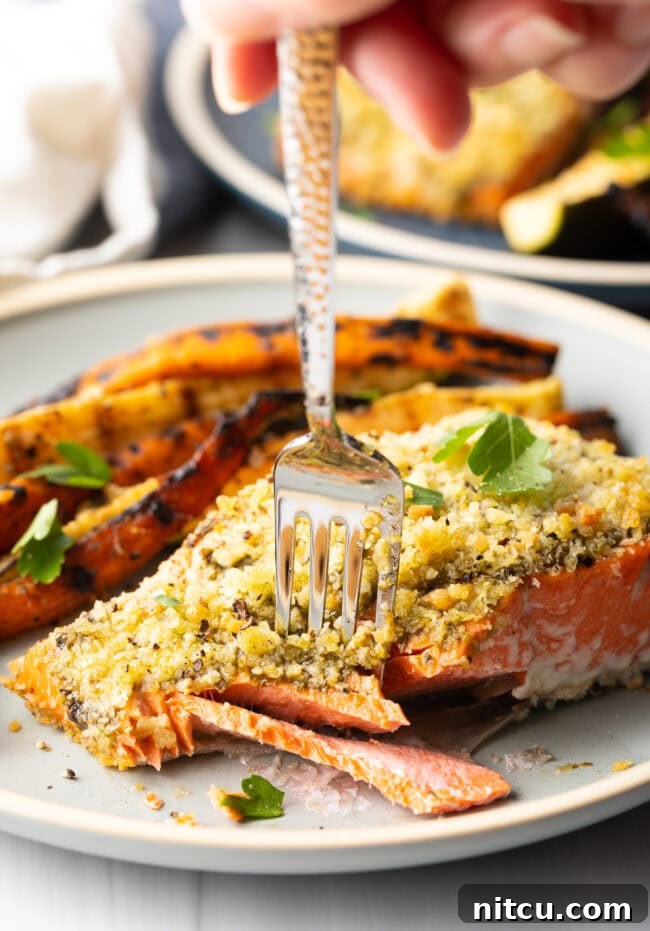 Fork getting a bite of flaky salmon with pesto from a plate, next to grilled veggies.
