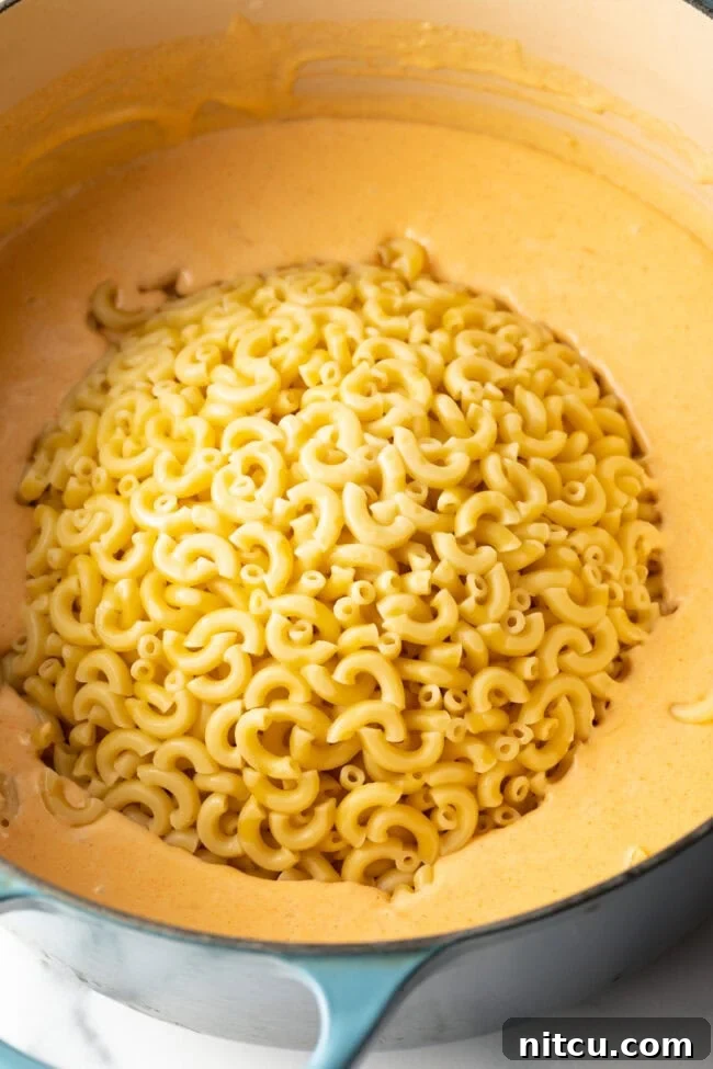 Drained elbow macaroni noodles being added to the pot with the rich cheese sauce.