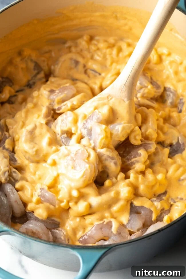 Wooden spoon stirring cheesy mac and cheese with uncooked shrimp in a large cooking pot.