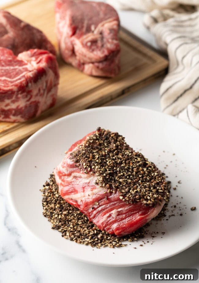 Uncooked steak being crusted in coarse black ground peppercorns on a plate.