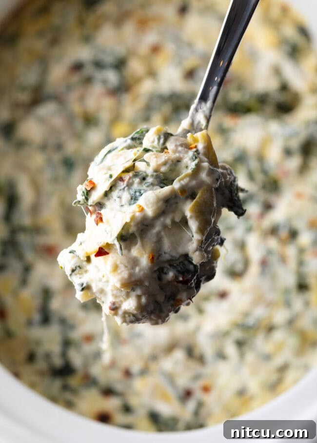 Spoon scooping slow cooker artichoke dip to camera.