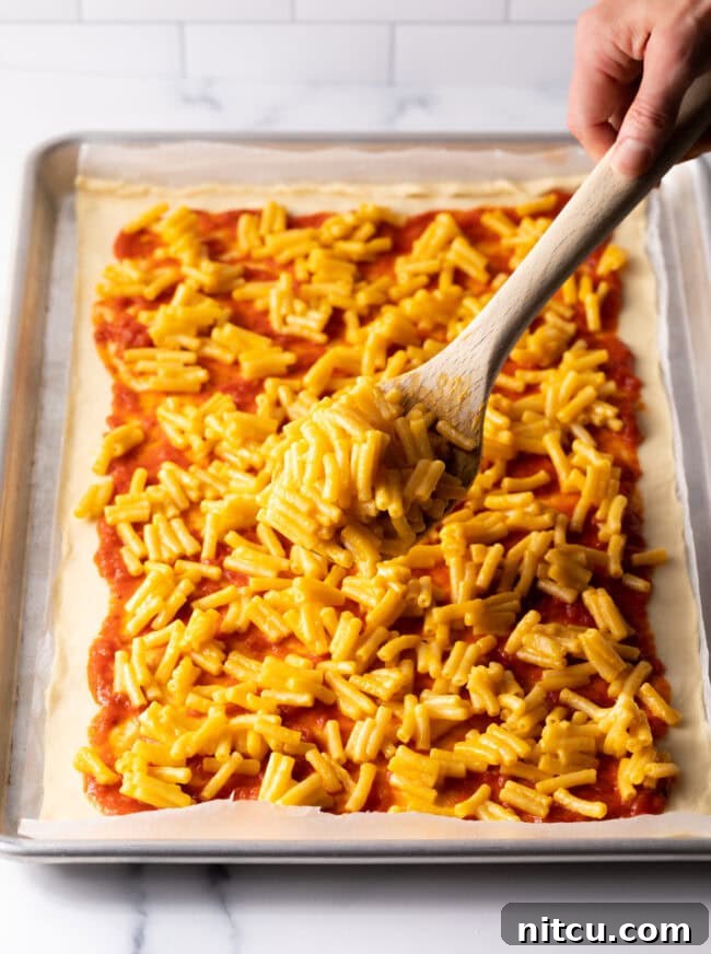 Cooked mac and cheese being spread over pizza sauce on dough.