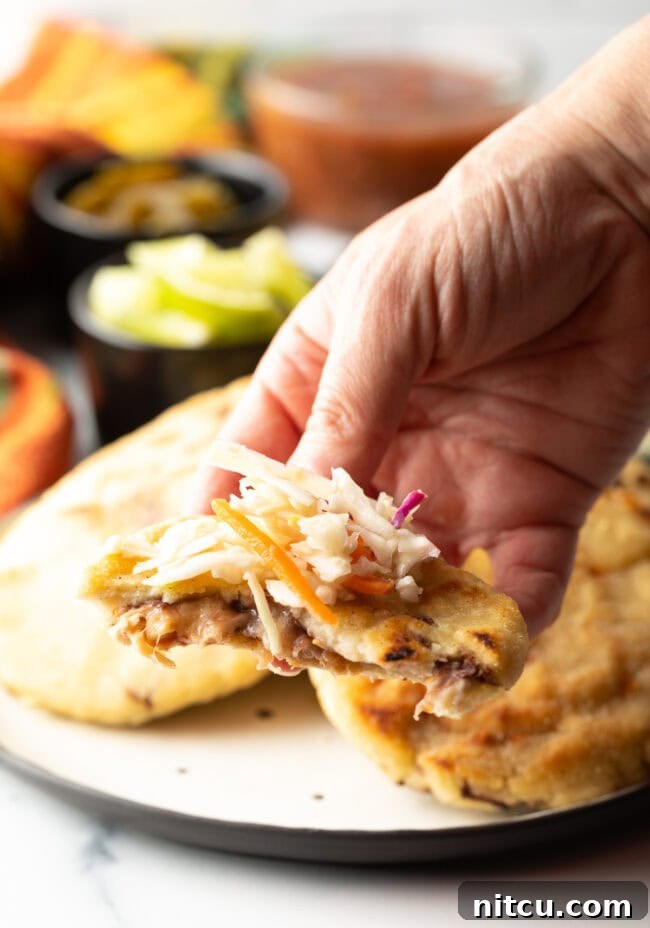 Hand holding a pupusa with a bite taken out, topped with curtido.